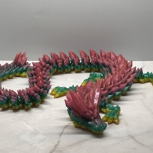May include: A colorful, articulated dragon figurine. The dragon is primarily pink, green, and yellow, with a crystalline texture. The body is segmented, allowing for posing. The dragon is lying on a white surface.