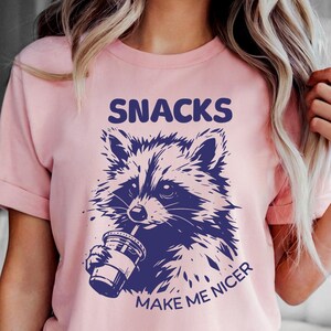 K&ouml;nnte beinhalten: Hellrosa T-Shirt mit einer dunkelblauen Waschb&auml;r-Grafik, die aus einem Becher trinkt. Der Text "SNACKS MAKE ME NICER" befindet sich &uuml;ber und unter der Waschb&auml;r-Illustration. Das Shirt wird mit Jeansshorts getragen.