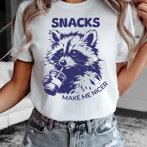 K&ouml;nnte beinhalten: Wei&szlig;es T-Shirt mit einem marineblauen Grafik eines Waschb&auml;ren, der aus einem Becher trinkt. Der Text "SNACKS MAKE ME NICER" befindet sich &uuml;ber und unter der Illustration. Das T-Shirt hat kurze &Auml;rmel.