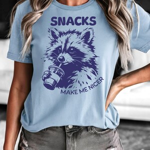 K&ouml;nnte beinhalten: Hellblaues T-Shirt mit einer lila Waschb&auml;r-Illustration, die aus einem Becher trinkt. Der Text "SNACKS MAKE ME NICER" befindet sich &uuml;ber und unter der Grafik. Ein l&auml;ssiges Kleidungsst&uuml;ck.