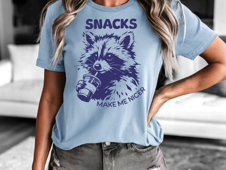 K&ouml;nnte beinhalten: Hellblaues T-Shirt mit einer lila Waschb&auml;r-Illustration, die aus einem Becher trinkt. Der Text "SNACKS MAKE ME NICER" befindet sich &uuml;ber und unter der Grafik. Ein l&auml;ssiges Kleidungsst&uuml;ck.