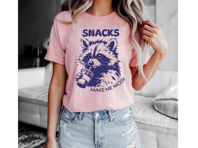 K&ouml;nnte beinhalten: Hellrosa T-Shirt mit einer dunkelblauen Waschb&auml;r-Grafik, die aus einem Becher trinkt. Der Text "SNACKS MAKE ME NICER" befindet sich &uuml;ber und unter der Waschb&auml;r-Illustration. Das Shirt wird mit Jeansshorts getragen.