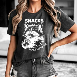 K&ouml;nnte beinhalten: Dunkelgraues T-Shirt mit einer wei&szlig;en Grafik eines Waschb&auml;ren, der ein Getr&auml;nk h&auml;lt. Der Text "SNACKS" steht &uuml;ber dem Waschb&auml;ren und "MAKE ME NICER" darunter. Das Shirt wird mit Jeansshorts getragen.