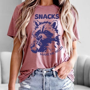 K&ouml;nnte beinhalten: Staubrosafarbenes T-Shirt mit einer Waschb&auml;r-Grafik, die aus einem Becher trinkt. Der marineblaue Text "SNACKS MAKE ME NICER" ist &uuml;ber und unter der Waschb&auml;r-Illustration aufgedruckt. Das Shirt ist aus weichem Material.