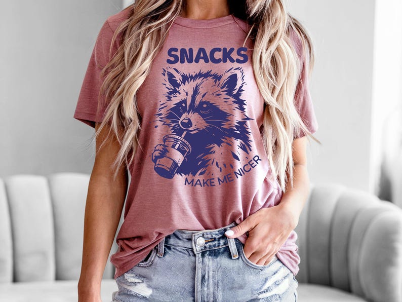 K&ouml;nnte beinhalten: Staubrosafarbenes T-Shirt mit einer Waschb&auml;r-Grafik, die aus einem Becher trinkt. Der marineblaue Text "SNACKS MAKE ME NICER" ist &uuml;ber und unter der Waschb&auml;r-Illustration aufgedruckt. Das Shirt ist aus weichem Material.