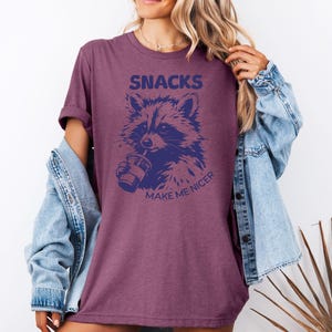 K&ouml;nnte beinhalten: Burgunderfarbenes T-Shirt mit einer marineblauen Waschb&auml;r-Grafik, die aus einem Becher trinkt. Der Text "SNACKS MAKE ME NICER" steht &uuml;ber und unter der Waschb&auml;r-Illustration. Das Shirt hat hochgekrempelte &Auml;rmel.