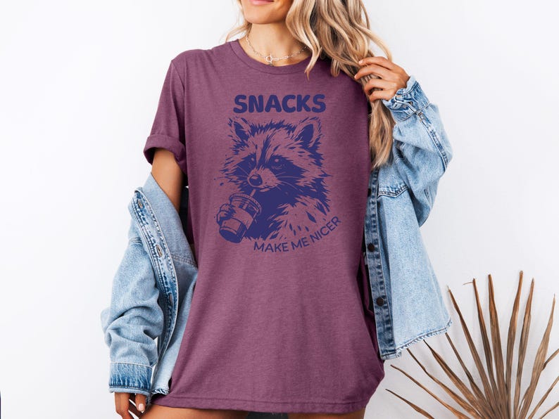 K&ouml;nnte beinhalten: Burgunderfarbenes T-Shirt mit einer marineblauen Waschb&auml;r-Grafik, die aus einem Becher trinkt. Der Text "SNACKS MAKE ME NICER" steht &uuml;ber und unter der Waschb&auml;r-Illustration. Das Shirt hat hochgekrempelte &Auml;rmel.