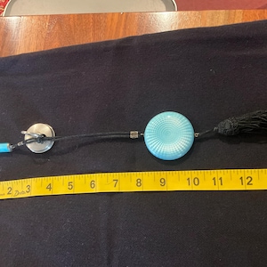May include: A light blue circular pendant with a radial pattern, attached to a black cord with a black tassel, a silver-toned metal ring, and a small light blue cylindrical charm. A yellow measuring tape shows the item is 14 inches in length.