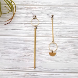 May include: A pair of gold earrings with geometric shapes. One earring features a long, thin rod with a circle at the top. The other earring has a long, thin rod with a circle and a half-circle at the bottom.