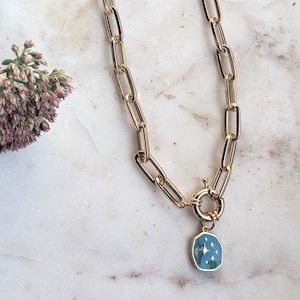 May include: Gold-toned chain necklace with a rectangular link design. The necklace features a pendant with a blue enamel background and a star design. The necklace is displayed on a white marble surface.