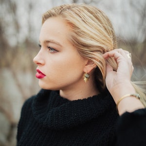 Pu&ograve; includere: Una donna con i capelli biondi indossa un maglione nero a collo alto e orecchini d'oro. Sta guardando da un'altra parte.