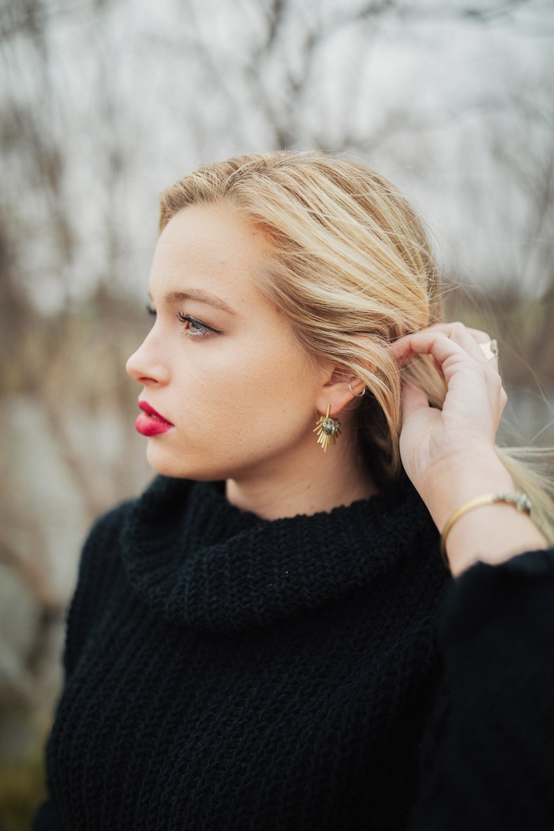 Pu&ograve; includere: Una donna con i capelli biondi indossa un maglione nero a collo alto e orecchini d'oro. Sta guardando da un'altra parte.