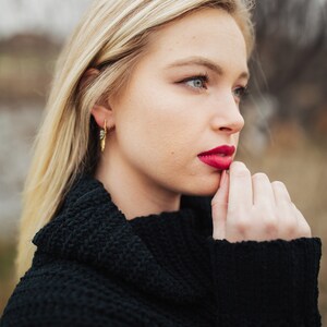 Pu&ograve; includere: Una donna con lunghi capelli biondi indossa un maglione nero a collo alto. Sta guardando da un lato e ha la mano sul mento. Indossa un rossetto rosso.
