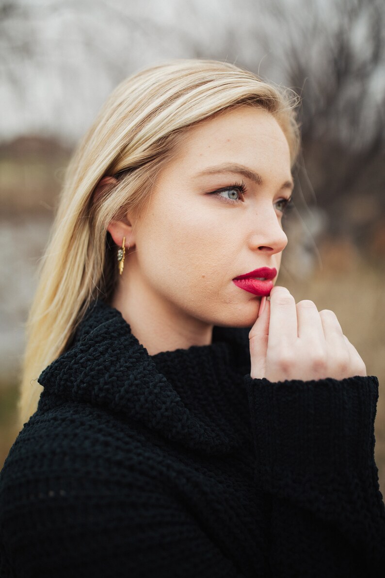 Pu&ograve; includere: Una donna con lunghi capelli biondi indossa un maglione nero a collo alto. Sta guardando da un lato e ha la mano sul mento. Indossa un rossetto rosso.