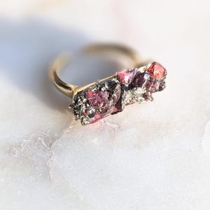 May include: A gold-toned ring featuring a cluster of raw, uncut gemstones in shades of pink and red. The gemstones are set on a textured, dark-colored base, creating a unique and organic design. The ring is set against a white marble surface.