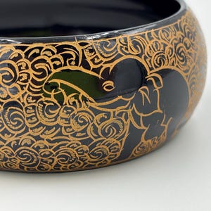 May include: Black bangle bracelet with a gold design of an elephant and swirling patterns.