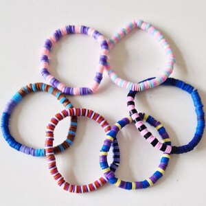 Op de afbeelding: Een verzameling kleurrijke kralenarmbanden in verschillende tinten blauw, paars, roze, bruin, rood, geel en zwart. De armbanden zijn in een cirkelvorm gerangschikt en tonen hun levendige en diverse kleurenschema's. Elke armband is gemaakt van kleine, platte kralen.