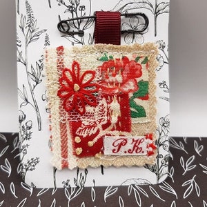 May include: A red and white fabric pin with a floral design and the initials "P.K." on a white background with black floral patterns.