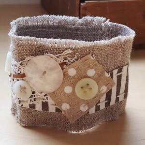 Op de afbeelding: Een bruin en wit stoffen armband met een witte knoop en een bruin en wit gestreept patroon. De armband is versierd met witte kant en een bruine knoop.