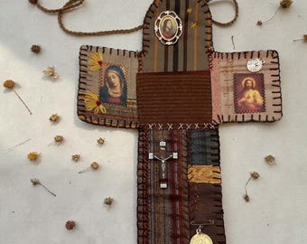 May include: Handmade cross-shaped religious item with fabric patches in brown, tan, and patterned textiles. Features embroidered details, religious imagery, and a small crucifix. Includes a cord for hanging and a gold-coloured medal.