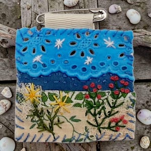 Peut inclure: Broche textile faite à la main représentant un paysage. La partie supérieure présente un motif bleu ajouré avec des accents blancs en forme d'étoile. En dessous, une section bleue texturée représente la mer, et le bas présente des fleurs et des plantes brodées. Une épingle de sûreté est attachée.