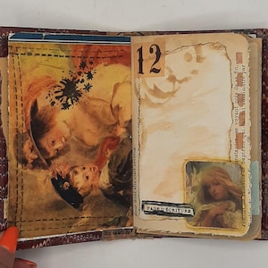 May include: Open journal with vintage-style pages. One page features a detailed illustration of children, while the facing page has the number 12 and a small image. The journal is bound with a brown ribbon.