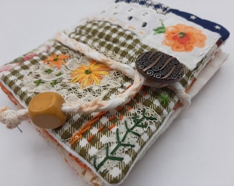 Puede incluir: Un pequeño diario de tela hecho a mano con una cubierta de patchwork verde, blanco y naranja. El diario está atado con un cordón blanco y tiene una cuenta de madera y un botón de metal.