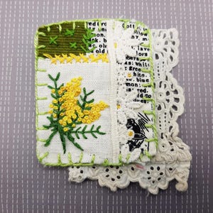 May include: A handmade textile art piece featuring embroidered yellow flowers and green leaves on white fabric. The piece is bordered with green stitching and includes a section of printed text and white lace trim. The background is a light gray with a dotted pattern.