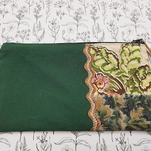 Peut inclure: Une pochette zippée en toile verte avec un rabat en tissu floral. Le rabat a un bord festonné et est décoré d'un motif floral brun et vert. La pochette a un tirette de fermeture éclair en cuir brun avec une perle verte.