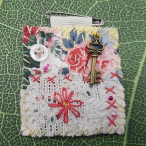 Puede incluir: Broche de tela hecha a mano con estampado floral, encaje y costuras decorativas. Un dije de llave de estilo vintage y un botón blanco están adjuntos. El broche se asegura con un imperdible.