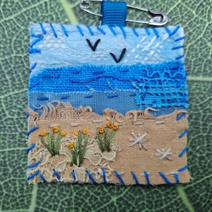 Può includere: Una toppa in tessuto quadrata che raffigura un paesaggio costiero con cielo blu, onde del mare e una spiaggia di sabbia beige ricamati. La scena include piccoli fiori gialli, steli verdi e due uccelli neri. È fissato un passante in nastro blu con una spilla.