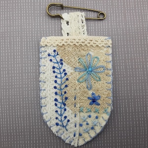 May include: Handmade, embroidered fabric pin with a decorative floral design. The pin features a cream-colored lace top, a beige and white patterned body, and a brass safety pin. Blue embroidered floral details adorn the fabric.