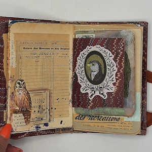 May include: Open vintage journal with aged paper pages. Features an owl sticker, a bird portrait in a wreath, and French text. The journal has a marbled burgundy cover and a brown ribbon closure. The pages have handwritten notes and stamps.