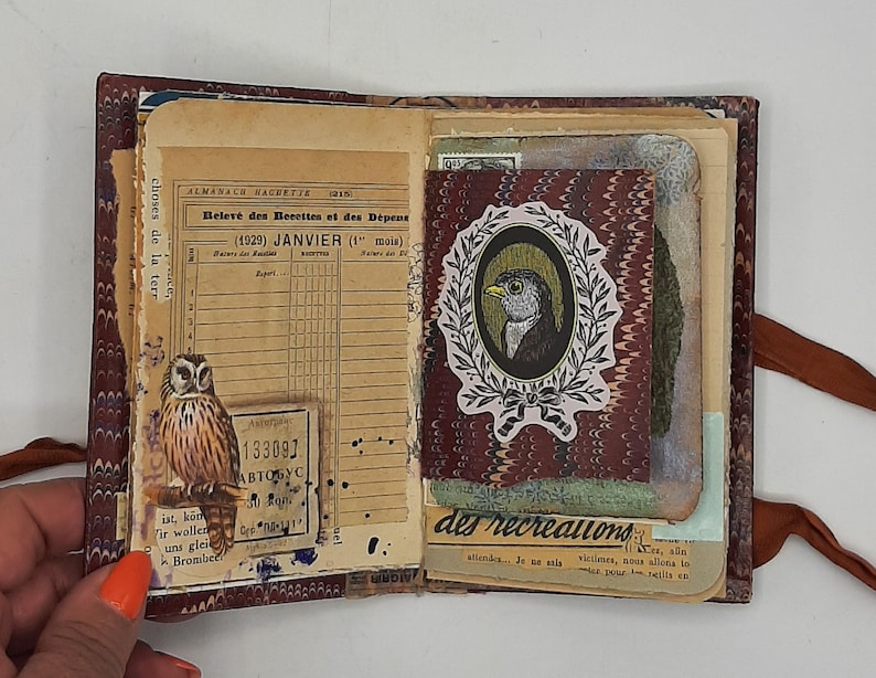 May include: Open vintage journal with aged paper pages. Features an owl sticker, a bird portrait in a wreath, and French text. The journal has a marbled burgundy cover and a brown ribbon closure. The pages have handwritten notes and stamps.