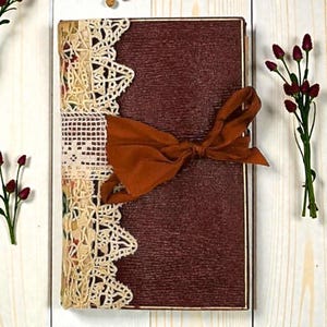 May include: A handmade journal with a burgundy cover and a decorative lace and ribbon closure. The journal is wrapped with a wide, rust-colored ribbon tied in a bow. The edges of the journal are adorned with cream-colored lace.