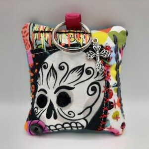 Peut inclure: Un porte-clés en tissu coloré avec un motif de crâne noir et blanc et un charme en croix argentée. Le porte-clés est fait de tissu patchwork avec une variété de couleurs et de motifs.