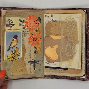 May include: Open journal with decorated pages. Features include a bird illustration, orange paper flowers, and a dragonfly drawing. The journal has a marbled cover and a brown ribbon closure. The pages have a vintage aesthetic.