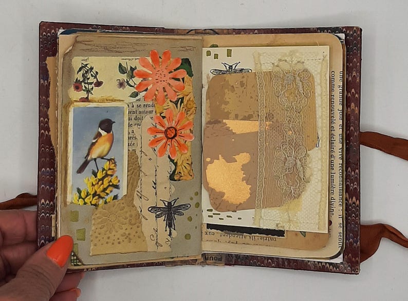 May include: Open journal with decorated pages. Features include a bird illustration, orange paper flowers, and a dragonfly drawing. The journal has a marbled cover and a brown ribbon closure. The pages have a vintage aesthetic.