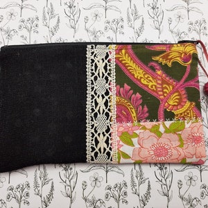 Peut inclure: Une pochette en tissu noir avec une fermeture éclair. La pochette est décorée d'un patchwork de tissus différents, dont une bordure en dentelle blanche, un imprimé paisley et un imprimé floral. La pochette a un pompon rouge.