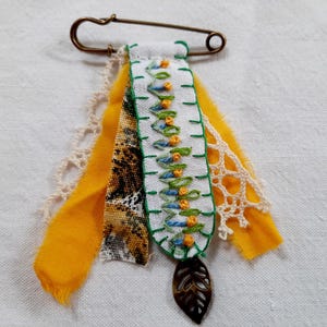 May include: A white fabric pin with green and blue embroidery, yellow and brown fabric accents, and a bronze leaf charm. The pin is attached to a safety pin.