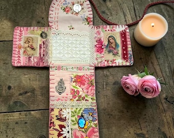 May include: A cross-shaped fabric wall hanging featuring patchwork floral patterns in pink, yellow, and green. It includes white lace trim, hand-stitched details, religious imagery, and small metal charms attached to the surface of the textile.