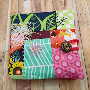 Peut inclure: Un objet en tissu patchwork coloré avec une fermeture à bouton. Le motif intègre divers motifs, notamment floraux, de feuilles et géométriques, dans des tons de vert, rose, orange et bleu. L'objet est orné de broderies et de dentelles.