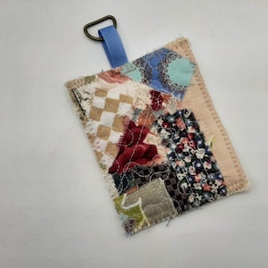 Può includere: Un piccolo portachiavi in tessuto patchwork con un anello di nastro blu. Il tessuto è un mix di motivi floreali, a quadri e geometrici in tonalità di marrone, rosso, blu e bianco.