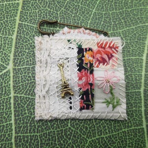 May include: A handmade fabric brooch featuring a patchwork design with floral patterns, lace, and a gold-colored Eiffel Tower charm. The brooch is secured with a safety pin. The color palette includes white, pink, black, and green.