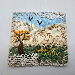 Peut inclure: Un patch en tissu carré représentant un paysage avec un ciel bleu clair, deux oiseaux noirs brodés, un arbre marron à nœuds jaunes, un sol vert avec des fleurs jaunes et des collines beiges, bordé par un point de croix beige décoratif.