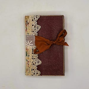 May include: A burgundy journal with a textured cover, gold trim, and a rust-colored ribbon tied in a bow. The left side is adorned with cream-colored lace and patterned fabric.