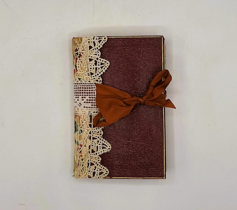 May include: A burgundy journal with a textured cover, gold trim, and a rust-colored ribbon tied in a bow. The left side is adorned with cream-colored lace and patterned fabric.