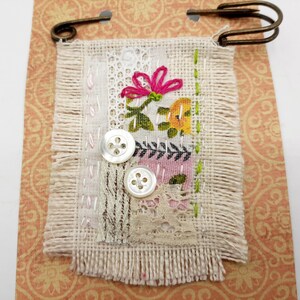 May include: A small, handmade fabric pin with a floral design. The pin is made of various fabrics, including lace, and features a pink flower, green leaves, and yellow and white buttons. The pin is attached to a metal pin back.