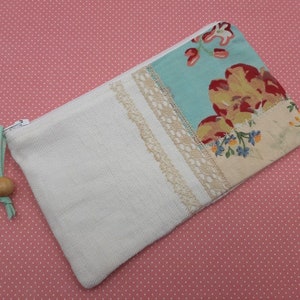 Op de afbeelding: Een witte ritszak met een klep van bloemenstof. De klep heeft een blauwe achtergrond met rode, gele en witte bloemen. De tas heeft een witte kantversiering en een rits met een houten kralen-trekker.