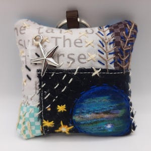 Peut inclure: Un petit porte-clés en tissu blanc avec un motif de planète bleu et vert, un charme d'étoile et le texte "The Universe" brodé dessus.